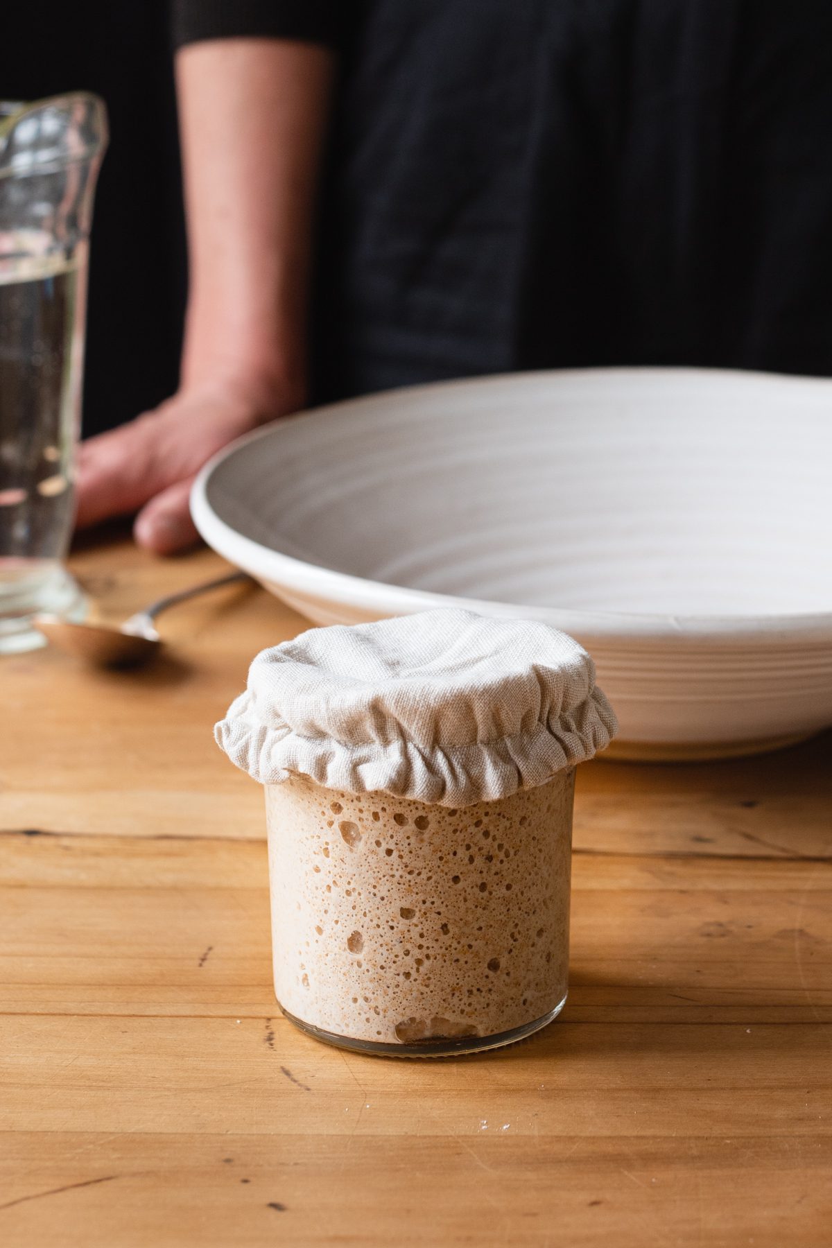 Jar of bubbly sourdough starter on kitchen bench. Mixing bowl, water and person in the background ready to make bread.